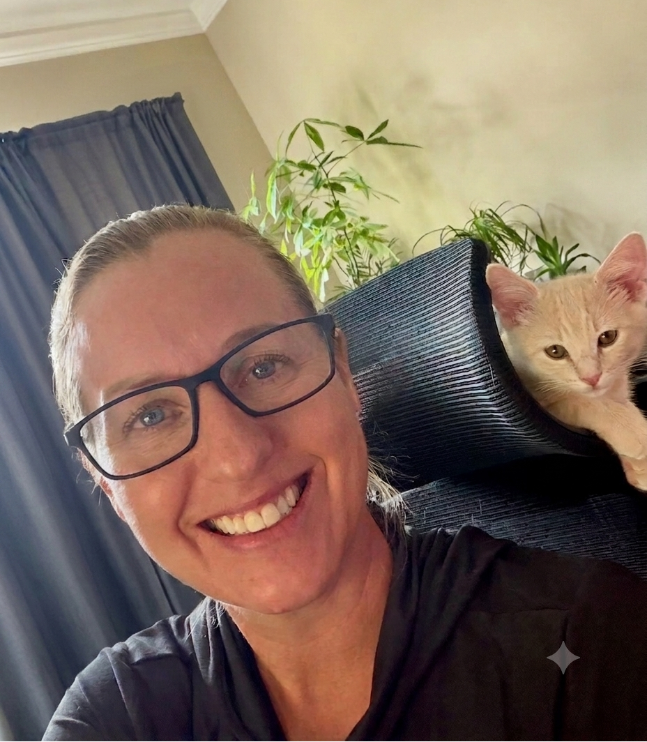 A smiling woman wearing black-rimmed glasses and a dark top takes a selfie indoors, with a light orange cat resting on a dark ribbed chair behind her and green plants in the background.